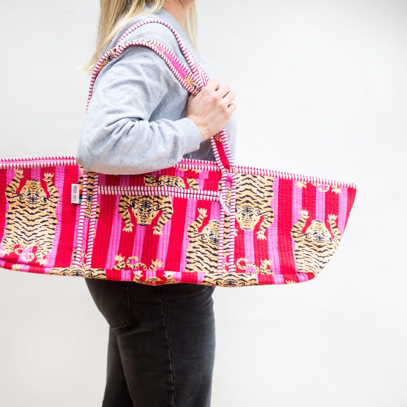 Puede incluir: Un bolso tote grande con un estampado de tigre vibrante. El bolso presenta un fondo a rayas rojas y rosas con caras de tigre. Las correas y los ribetes son rosas y blancos. El bolso lo sostiene una persona.