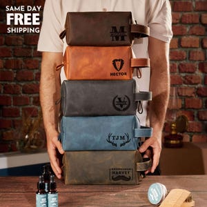 May include: A stack of five personalized leather toiletry bags in various colors: brown, orange, gray, blue, and tan. Each bag features a unique design, including initials, names, and a tuxedo graphic. The bags have a handle and zipper closure.
