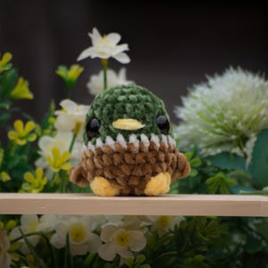 Op de afbeelding: Een klein gehaakt knuffel eendje met groene en bruine vacht, zwarte ogen en een oranje snavel. De eend zit op een houten plank voor een achtergrond van groene planten en witte bloemen.
