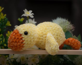 May include: A handmade, plush yellow and orange duck toy with a black eye. The duck is made of soft, textured yarn and has a rounded body, wings, and tail. The toy is resting on a wooden shelf, surrounded by flowers.