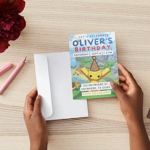 Op de afbeelding: Een verjaardagsuitnodiging met de tekst "LET'S CELEBRATE OLIVER'S BIRTHDAY" en evenementdetails. De uitnodiging toont een cartoonfiguur en wordt vastgehouden met een witte envelop. Een bordeauxrode bloem en roze pen liggen op het houten oppervlak.