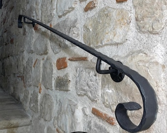 May include: A black hand-forged iron handrail featuring a textured surface and a decorative scrolled end. The rail is mounted to a wall with two circular brackets. Below the rail, text reads MADE IN ITALY FERMEG with a green, white, and red stripe.