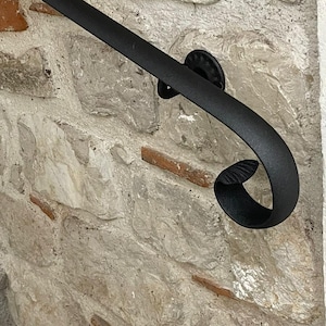 Puede incluir: Barandilla de hierro forjado negro con un extremo decorativo en forma de voluta, montada en una pared de piedra. La pared está construida con piedras de varios tamaños e intercalada con ladrillos rojos. La barandilla se curva.