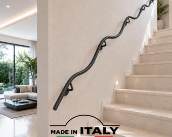 May include: A matte black metal handrail with a continuous wavy, serpentine design mounted to a light-colored wall alongside a staircase. The handrail features triangular support brackets and includes the text MADE IN ITALY FERMEG at the bottom.
