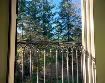 May include: A window with a wrought iron railing looking out onto a forest of evergreen trees. The window is open and the sun is shining through.