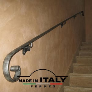 Peut inclure: Une rampe d'escalier en métal forgé à la main avec une finition gris argenté texturée et une extrémité décorative en volute. La rampe est fixée au mur par des supports métalliques et porte le texte MADE IN ITALY FERMEG avec un drapeau italien.