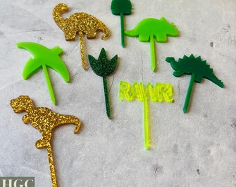 May include: A collection of dinosaur-themed cake toppers in various shades of green and gold glitter. The toppers include dinosaur silhouettes and the word "RAWR". The image also includes the text "SVG & PDF LASER FILE + COMMERCIAL USE".