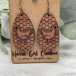 Könnte beinhalten: Ein Paar Holzohrringe mit einem Hühnerdesign auf braunem Hintergrund. Die Ohrringe sind oval geformt und haben ein Wabenmuster. Die Ohrringe hängen an einer braunen Karte mit dem Text "Harris Girl Creations a mother & daughter team".