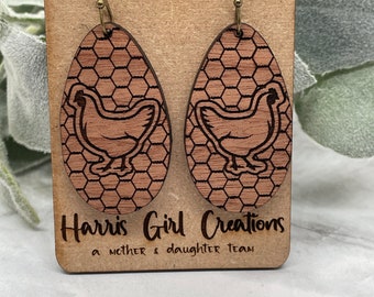 May include: A pair of wooden earrings with a chicken design on a brown background. The earrings are oval shaped and have a honeycomb pattern. The earrings are hanging on a brown card with the text "Harris Girl Creations a mother & daughter team".