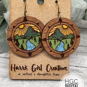 Puede incluir: Un par de pendientes de madera con una escena de la naturaleza. Los pendientes son redondos y presentan una montaña, un sol y un río. Los pendientes están en una tarjeta marrón con el texto "Harris Girl Creations a mother & daughter team" y "HGC DIGITAL DESIGNS".