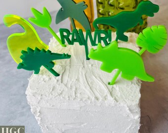 May include: A white frosted cake decorated with green and yellow dinosaur-themed toppers. The toppers include various dinosaur shapes and the word "RAWR!" in green. The cake is a fun and festive centerpiece.
