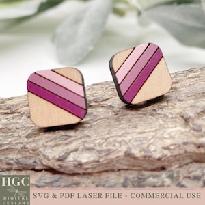 May include: A pair of square wooden earrings with a diagonal stripe pattern in shades of pink and purple. The earrings are shown on a wooden surface.
