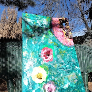 May include: Abstract floral painting on canvas. The artwork features a textured turquoise background with large, colorful flower shapes in pink, red, and yellow. The painting is displayed outdoors, leaning against a green wicker chair.