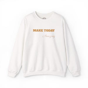 Puede incluir: Una sudadera blanca de manga larga con cuello redondo que presenta el texto "MAKE TODAY" en una fuente naranja, audaz y texturizada, seguido de la palabra "Amazing" en una fuente cursiva más pequeña y elegante en el pecho.