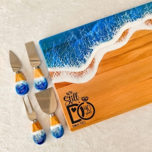 May include: A rectangular wooden serving board with a blue and white resin ocean wave design and four matching cheese knives. The board features black text that reads We Still DO 40 years since 1985 with a diamond ring graphic.