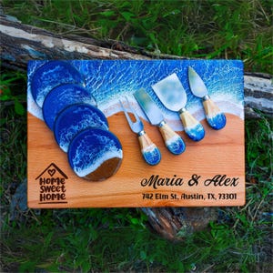 May include: Wooden serving board with a beach-themed design, featuring blue and white resin coasters and cheese knives. The board is engraved with "Home Sweet Home" and the names "Maria & Alex".