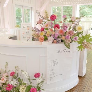 May include: A white bar with a curved front, decorated with a floral arrangement of pink, white, and purple flowers. A sign on the bar reads "Bar Menu" and lists various spirits, beer, wine, and assorted soft drinks.