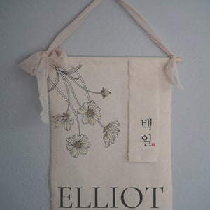 May include: A beige fabric banner with black floral illustrations and the name "ELLIOT" printed in black. The banner also features Korean text in black and red. The banner is hanging from a light pink ribbon.