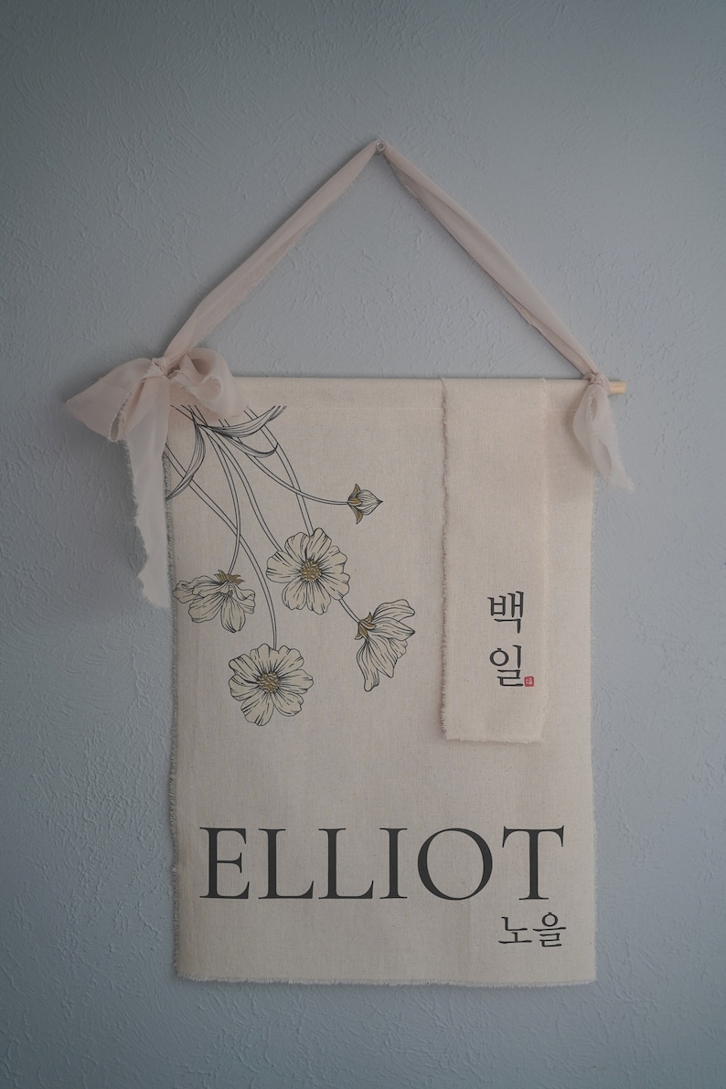 May include: A beige fabric banner with black floral illustrations and the name "ELLIOT" printed in black. The banner also features Korean text in black and red. The banner is hanging from a light pink ribbon.