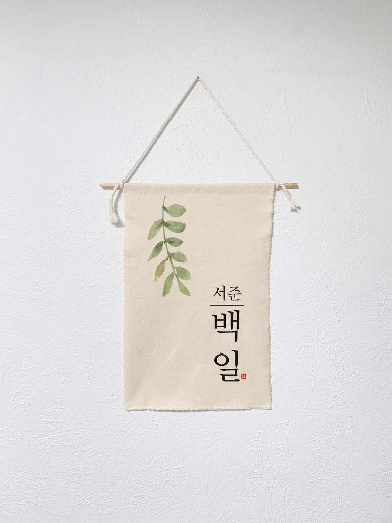 May include: A beige canvas wall hanging with a green watercolor leaf design and the Korean text "서른 밤" in black. The hanging is attached to a white wall with a wooden dowel and twine.