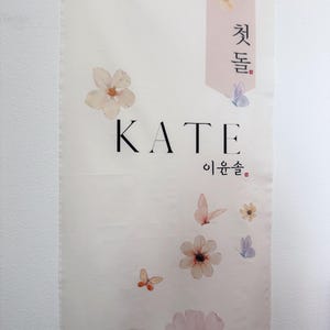 May include: A white banner with the name "KATE" in black lettering. The banner is decorated with delicate pink and white flowers and butterflies. The Korean text "첫돌" and "이환솔" are also on the banner.