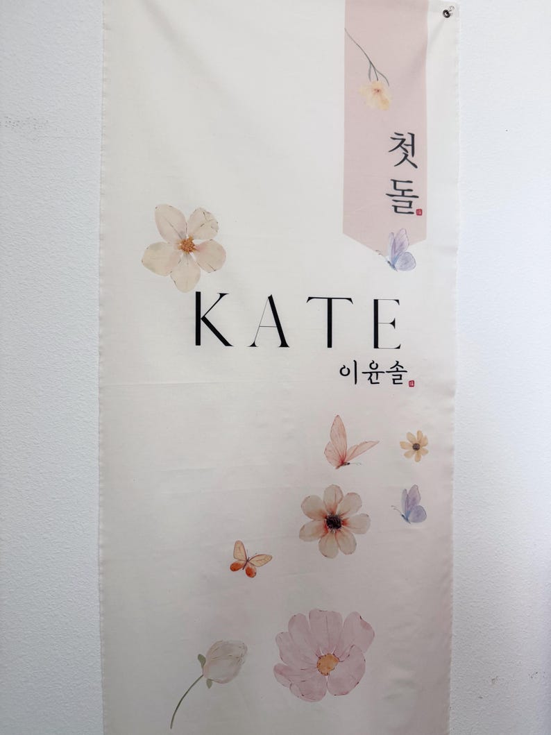 May include: A white banner with the name "KATE" in black lettering. The banner is decorated with delicate pink and white flowers and butterflies. The Korean text "첫돌" and "이환솔" are also on the banner.