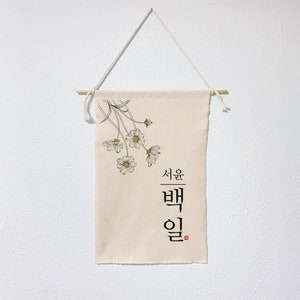 May include: A beige canvas wall hanging with a floral design and the Korean text "Seoul Baek-il" in black ink. The hanging is attached to a white wall with a wooden dowel and twine.