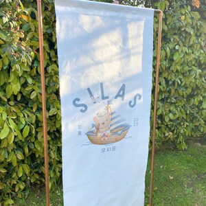 May include: A white canvas banner with the name Silas printed in blue and a cartoon bear wearing a sailor hat on a boat. The banner also includes the date 2023 and the Korean word for "sail" in blue.