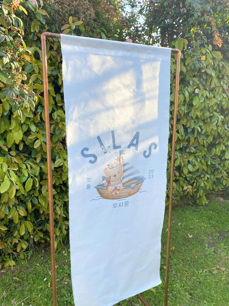 May include: A white canvas banner with the name Silas printed in blue and a cartoon bear wearing a sailor hat on a boat. The banner also includes the date 2023 and the Korean word for "sail" in blue.