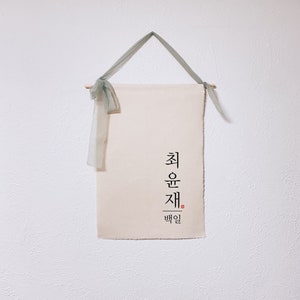 May include: A white canvas wall hanging with the Korean text "최운재" and the word "백일" in smaller font below. The hanging is attached to a wooden dowel with a green ribbon.