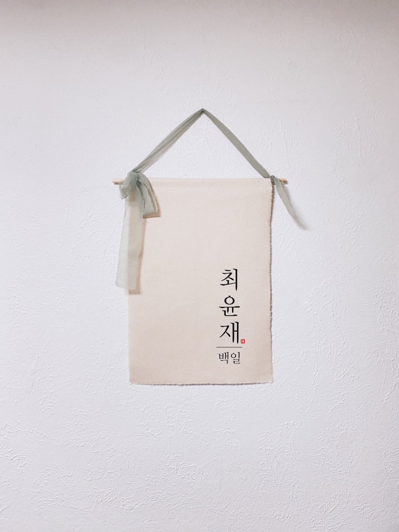 May include: A white canvas wall hanging with the Korean text "최운재" and the word "백일" in smaller font below. The hanging is attached to a wooden dowel with a green ribbon.