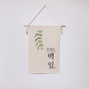 May include: A beige canvas banner with a green watercolor leaf design and the name "JOEL" written in black lettering. Below the name is the Korean word "백일" written in black calligraphy.