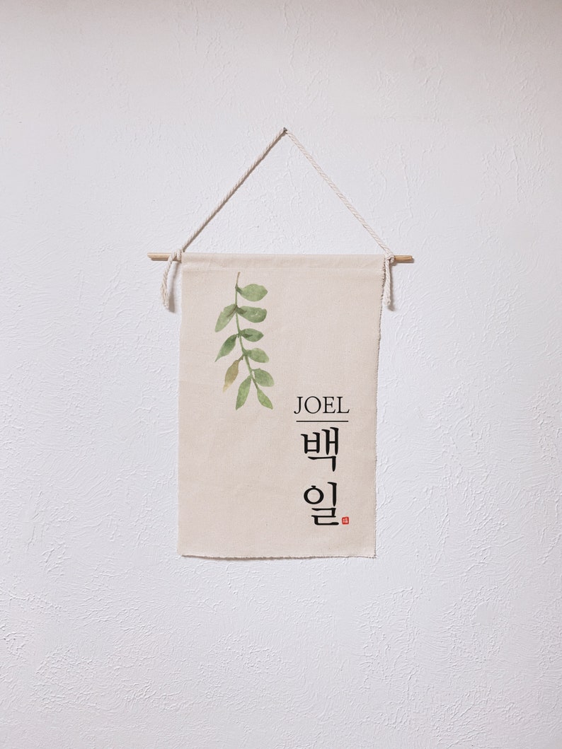 May include: A beige canvas banner with a green watercolor leaf design and the name "JOEL" written in black lettering. Below the name is the Korean word "백일" written in black calligraphy.