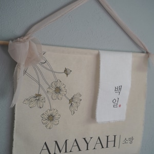 May include: A beige canvas banner with a floral design and the name "AMAYAH" in black lettering. The banner also includes the Korean text "뱁" and "소망" in black lettering.