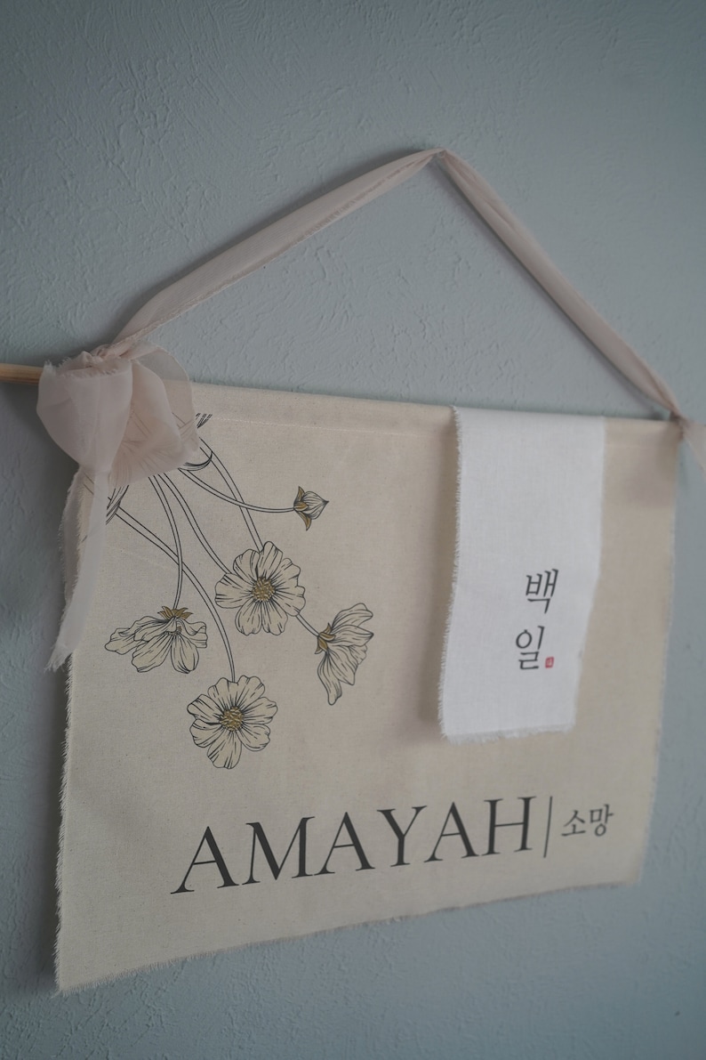 May include: A beige canvas banner with a floral design and the name "AMAYAH" in black lettering. The banner also includes the Korean text "뱁" and "소망" in black lettering.