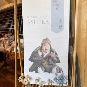 May include: A white banner with the text "Welcome to Asher's" and a photo of a baby in traditional Korean clothing. The banner is hanging on a wooden stand with small vases of flowers.