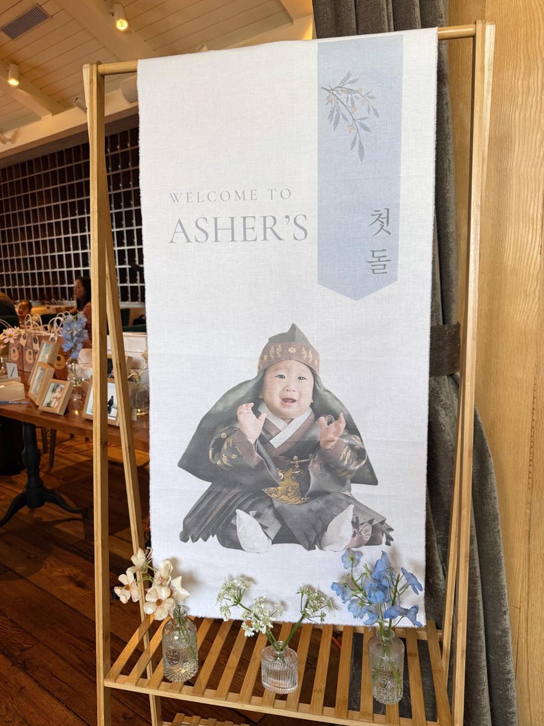 May include: A white banner with the text "Welcome to Asher's" and a photo of a baby in traditional Korean clothing. The banner is hanging on a wooden stand with small vases of flowers.