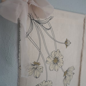 May include: A white canvas wall hanging with a floral design. The flowers are outlined in black and have a yellow center. A light pink ribbon is tied at the top of the hanging.
