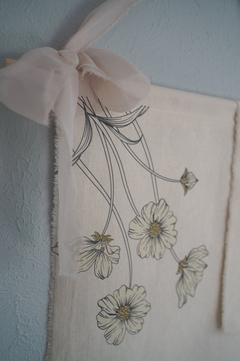 May include: A white canvas wall hanging with a floral design. The flowers are outlined in black and have a yellow center. A light pink ribbon is tied at the top of the hanging.
