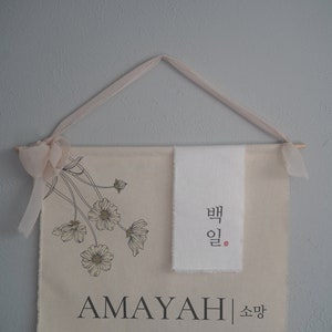 May include: A beige canvas wall hanging with a floral design and the name "AMAYAH" in black lettering. The hanging also features a white fabric panel with Korean text that reads "밤" and "일".