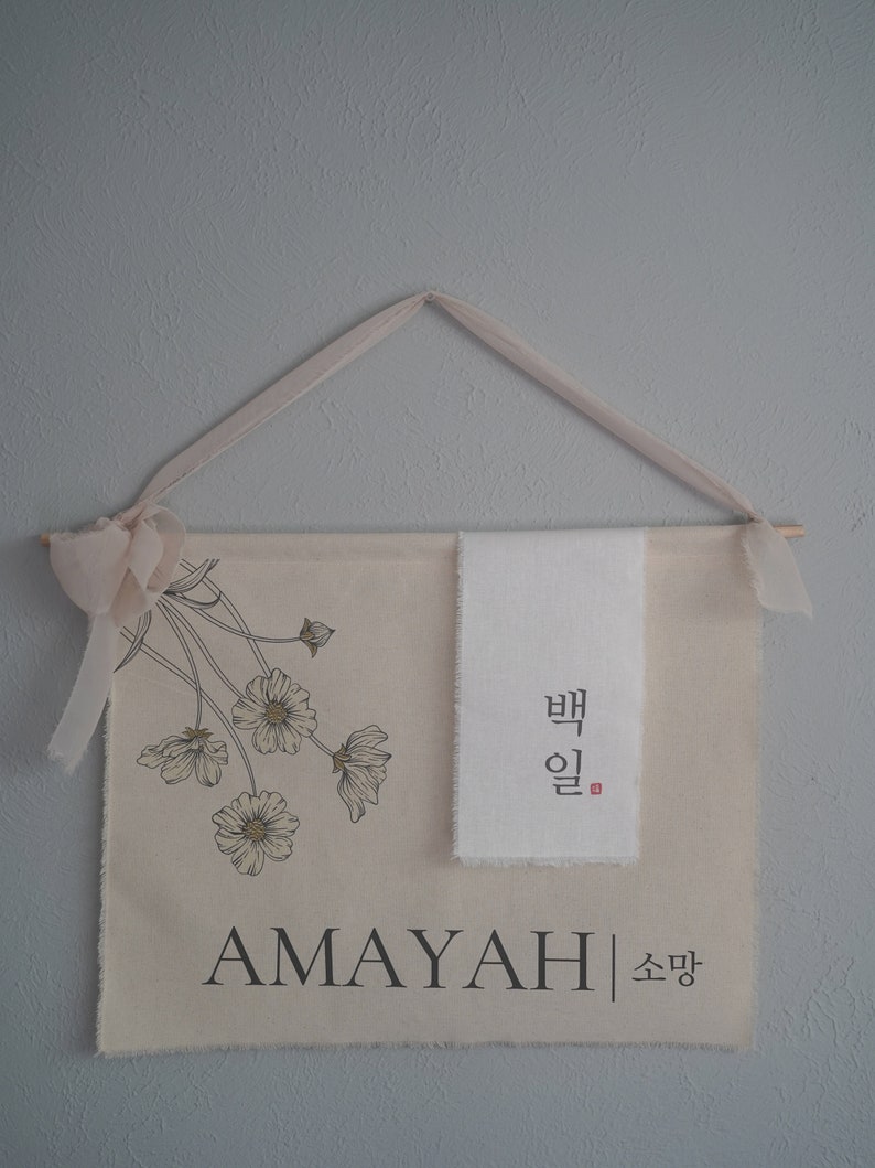 May include: A beige canvas wall hanging with a floral design and the name "AMAYAH" in black lettering. The hanging also features a white fabric panel with Korean text that reads "밤" and "일".