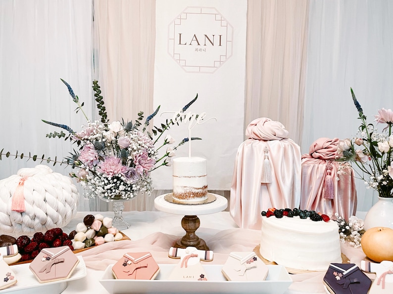 May include: A table decorated for a baby shower with a pink and white color scheme. The table features a white cake with a topper that says "Lani", a white cake with berries, and a variety of other treats. The table is decorated with pink and white flowers, and a sign that says "Lani" in Korean.