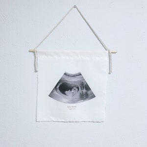 May include: A white fabric banner with a black and white ultrasound image of a baby. The banner has the text "Baby Peanut August 2023" printed on it.