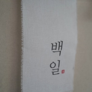 May include: A white fabric banner with the Korean words "백일" written in black ink. The words are centered on the banner and are surrounded by a white border.