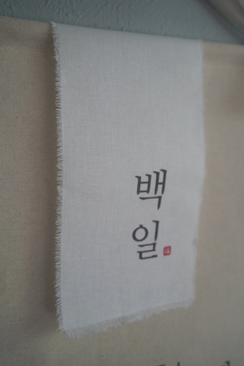 May include: A white fabric banner with the Korean words "백일" written in black ink. The words are centered on the banner and are surrounded by a white border.
