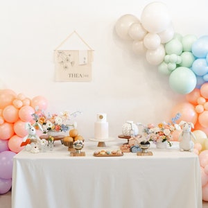May include: A white table with a white tablecloth is set up for a party. There are pastel balloons in the background, a white cake with a gold circle on top, and a sign that says "THEA 18e".