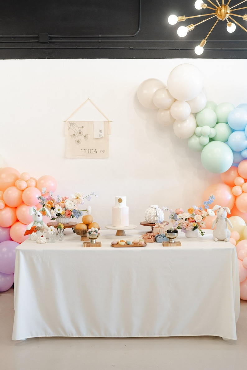 May include: A white table with a white tablecloth is set up for a party. There are pastel balloons in the background, a white cake with a gold circle on top, and a sign that says "THEA 18e".