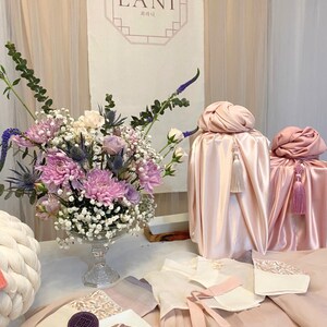 May include: A table setting with a pink and white theme. There are two pink silk-wrapped gifts with tassels, a bouquet of pink and white flowers in a clear glass vase, and a plate of cookies decorated with traditional Korean designs. The sign behind the table reads "LANI" in a decorative font.