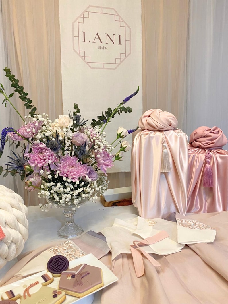May include: A table setting with a pink and white theme. There are two pink silk-wrapped gifts with tassels, a bouquet of pink and white flowers in a clear glass vase, and a plate of cookies decorated with traditional Korean designs. The sign behind the table reads "LANI" in a decorative font.