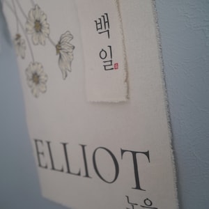 May include: A white canvas banner with the name "ELLIOT" printed in black. Above the name is a smaller banner with the Korean word "Elliot" written in black with a red seal. The banner is decorated with a floral design.